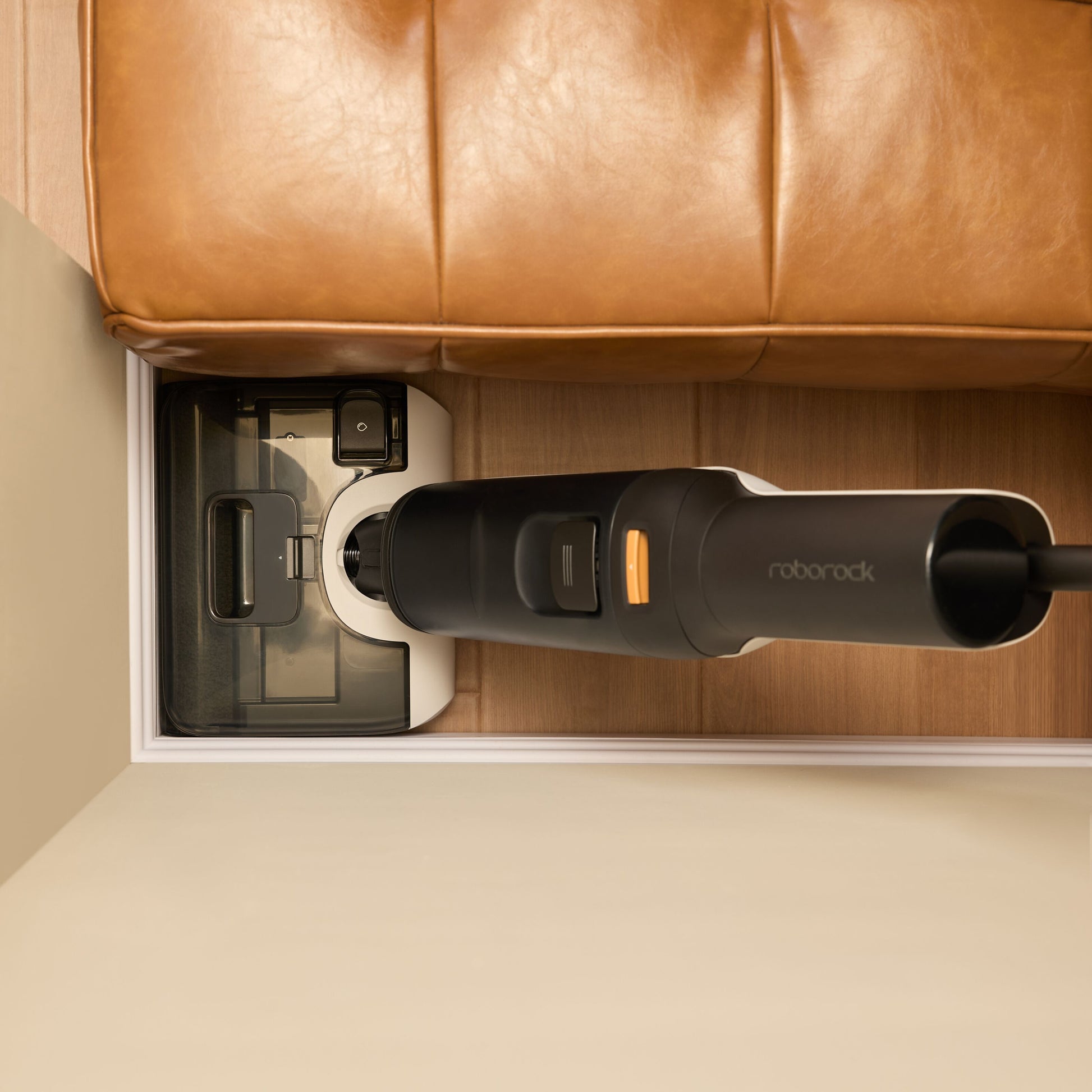 Roborock F25 Støvsuger Sort i ladestation ved brun lædersofa på trægulv, lav højde giver tæt pasform under sofa.