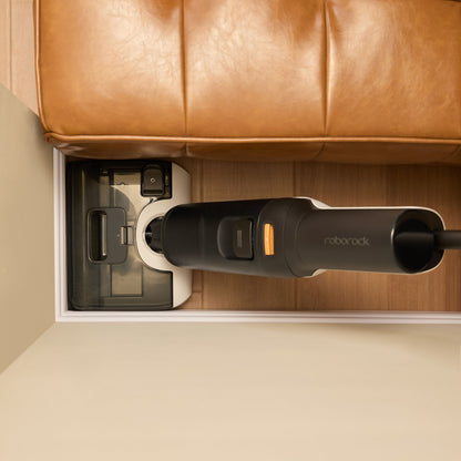 Roborock F25 Støvsuger Sort i ladestation ved brun lædersofa på trægulv, lav højde giver tæt pasform under sofa.