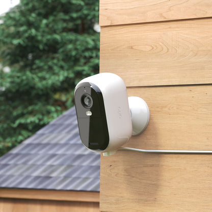 Arlo Essential 2 udendørskabel monteret på trævæg, synlige træer og hustag i baggrunden.