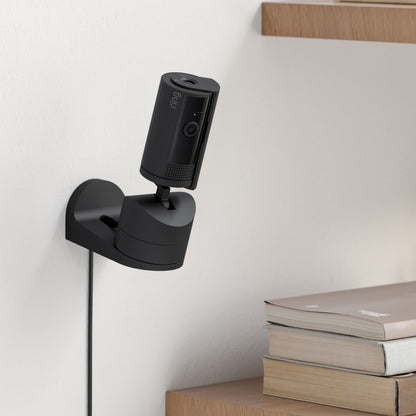 Ring Pan-Tilt indendørs kamera sort 1080p HD monteret på hvid væg nær træhylde med bøger strømtilslutning og kabel synligt.