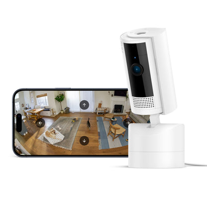 Ring Pan-Tilt indendørs kamera hvid ved smartphone med live-feed fra stue med tæppe og hund.
