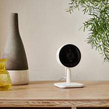 Yale Smart indendørskamera hvid på træbord ved grå-hvid vase, gult glas og grøn plante.