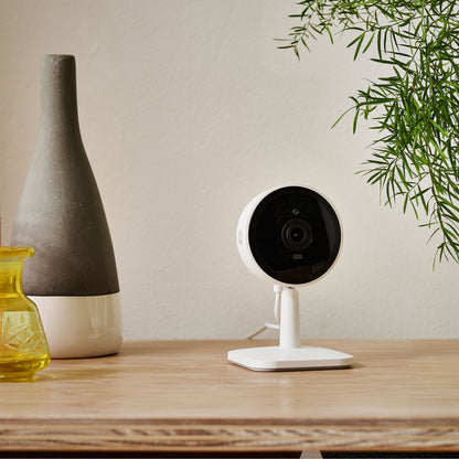 Yale Smart indendørskamera hvid på træbord ved grå-hvid vase, gult glas og grøn plante.