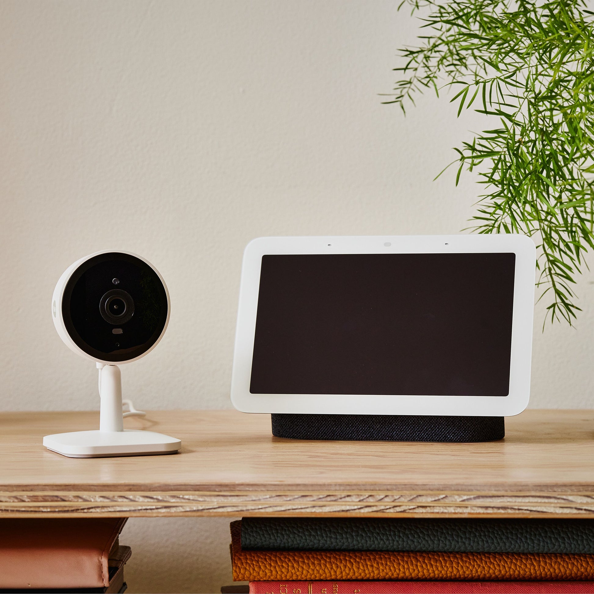 Yale Smart indendørskamera på træhylde ved siden af smart display med blank skærm, bøger og grøn plante.