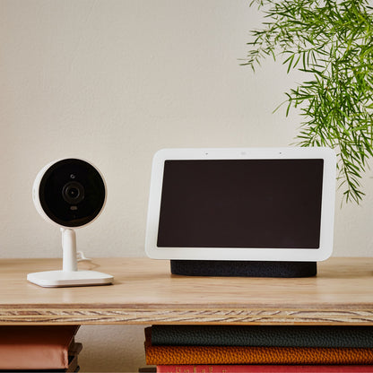 Yale Smart indendørskamera på træhylde ved siden af smart display med blank skærm, bøger og grøn plante.