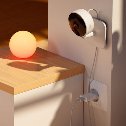 Yale Smart indendørskamera vægmonteret med strømtilslutning, orange kuglelampe placeres på bord ved siden af.