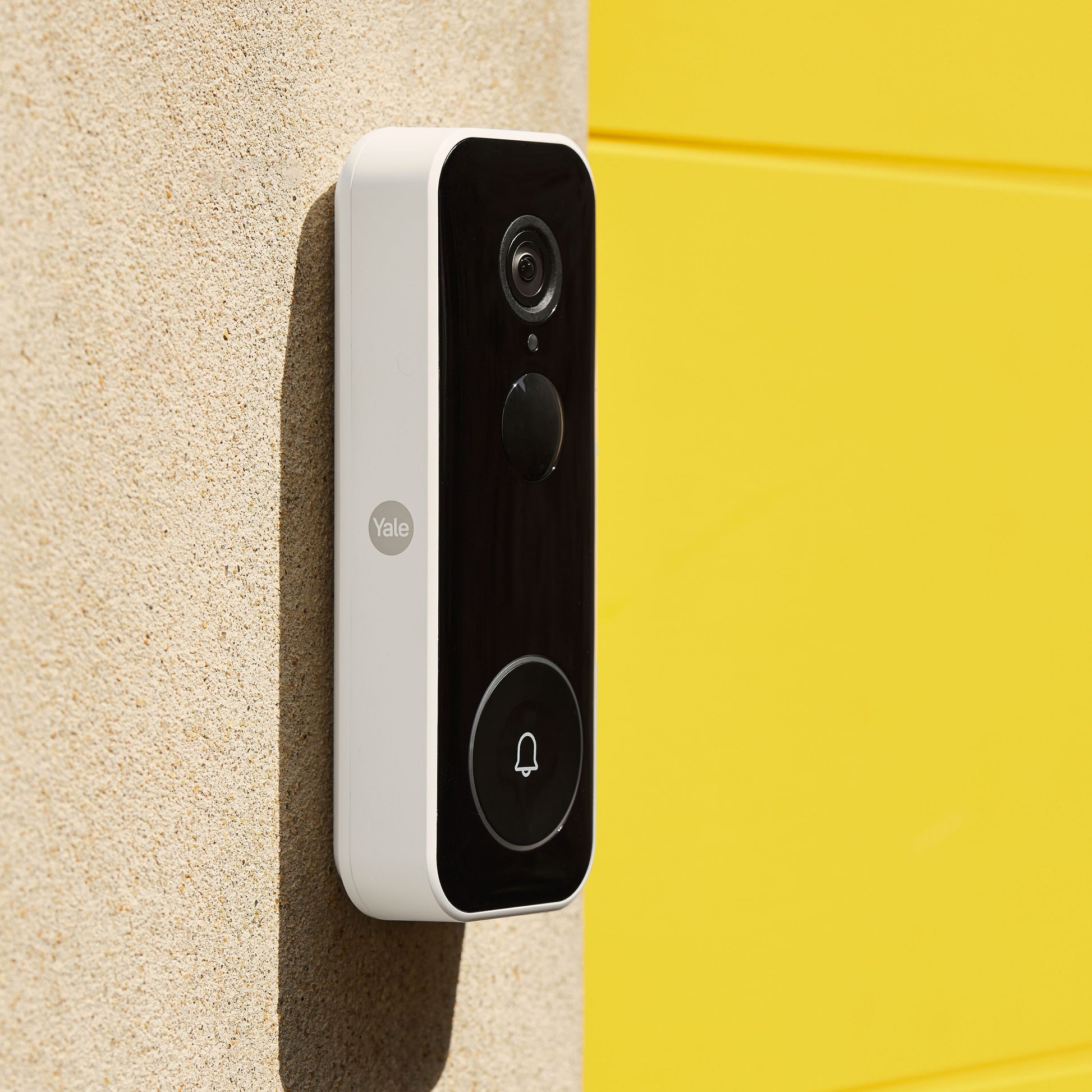 Yale Smart video dørklokke sort frontpanel hvide sider monteret på beige væg ved gul flade.