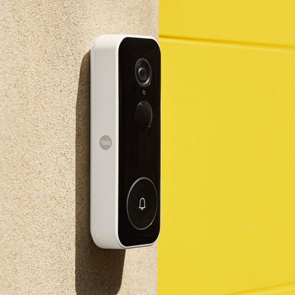 Yale Smart video dørklokke sort frontpanel hvide sider monteret på beige væg ved gul flade.