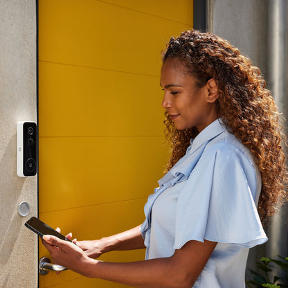 Yale Smart video dørklokke ved gul dør, kvinde holder smartphone nær enheden, smart home adgang.