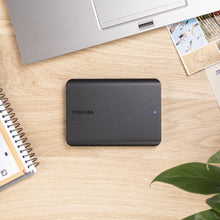Toshiba Canvio Basics ekstern harddisk 4TB med blå LED-lys på bord ved siden af bærbar, notesbog og fotos.
