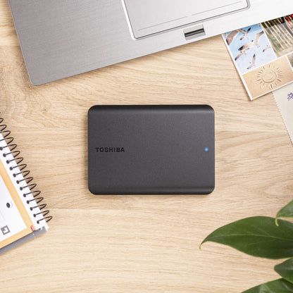 Toshiba Canvio Basics ekstern harddisk 4TB med blå LED-lys på bord ved siden af bærbar, notesbog og fotos.
