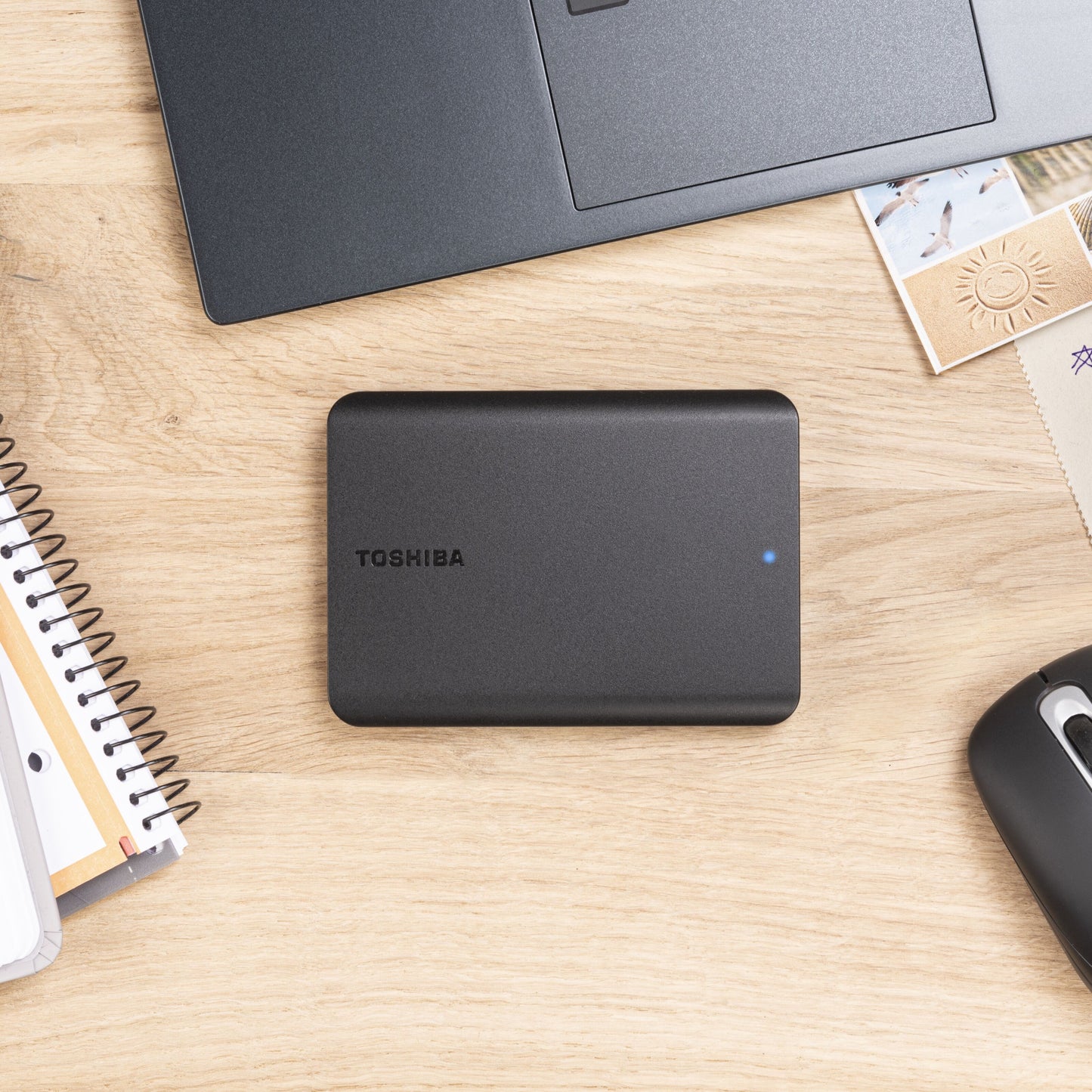 Toshiba Canvio Basics ekstern harddisk 4TB sort med blå indikatorlys ved siden af laptop, notesbog, mus og kuverter.