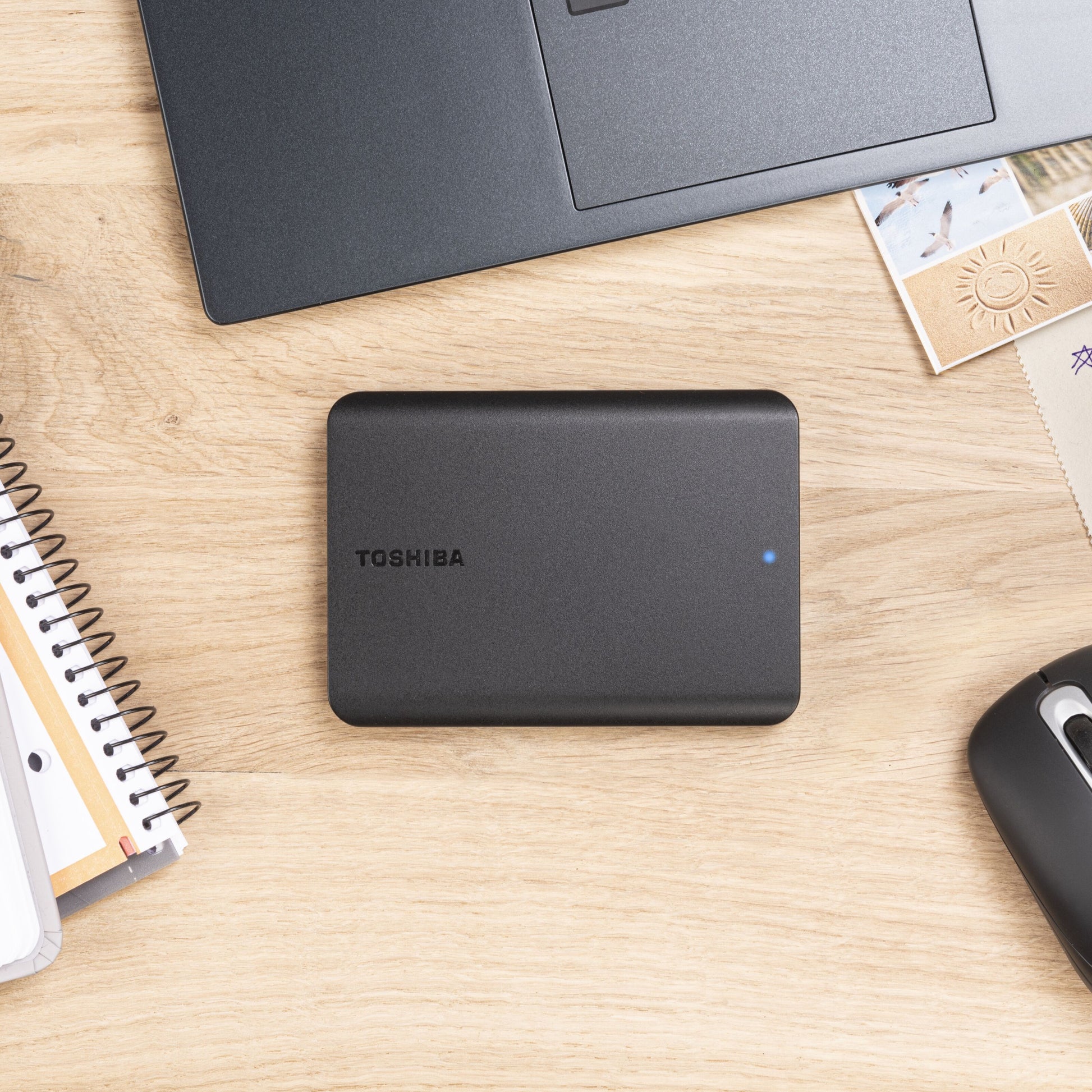 Toshiba Canvio Basics ekstern harddisk 4TB sort med blå indikatorlys ved siden af laptop, notesbog, mus og kuverter.