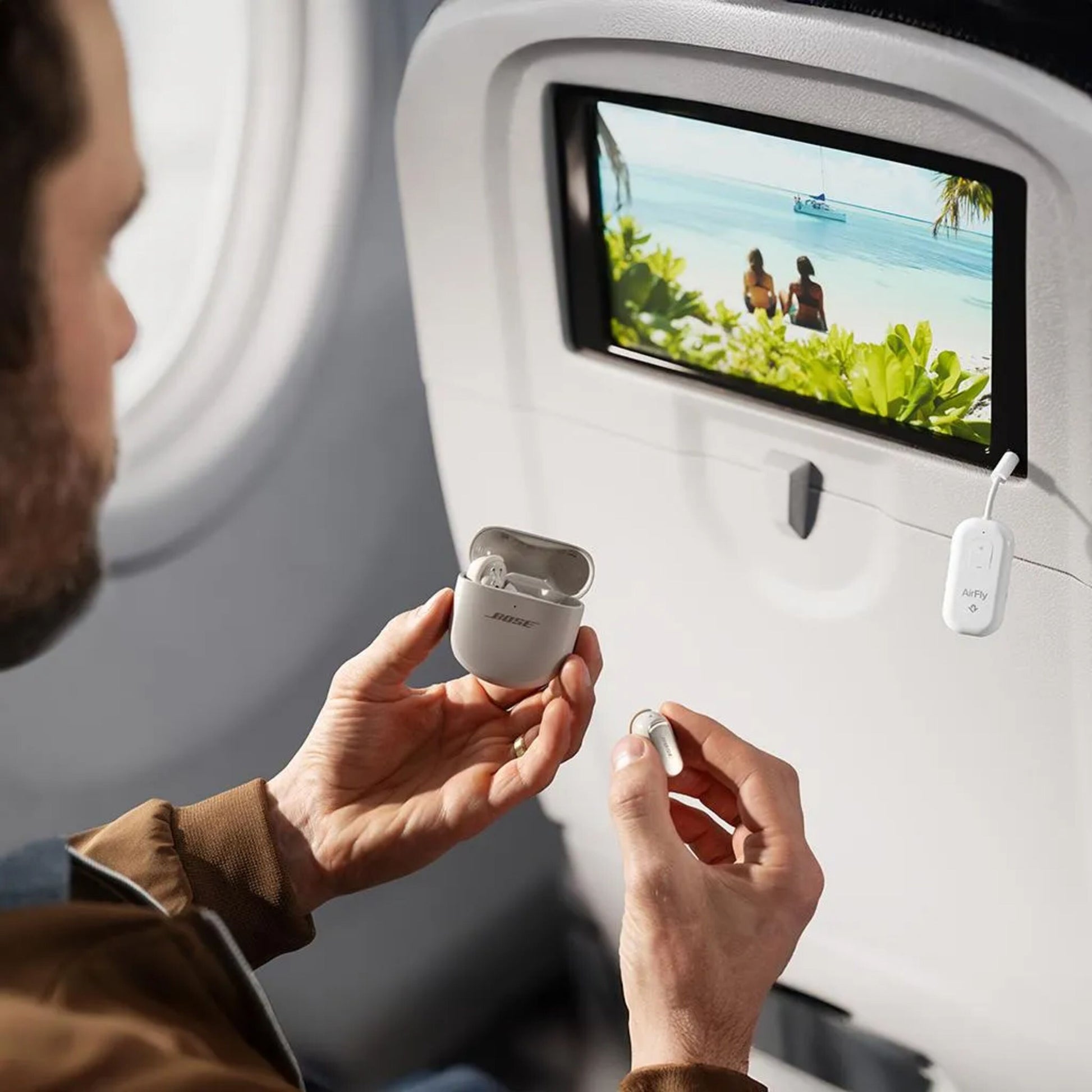 AirFly Pro 2 Bluetooth-sender og Bose-earbuds i hånd på fly, lydadapter og sædeskærm med strandmotiv synlige.