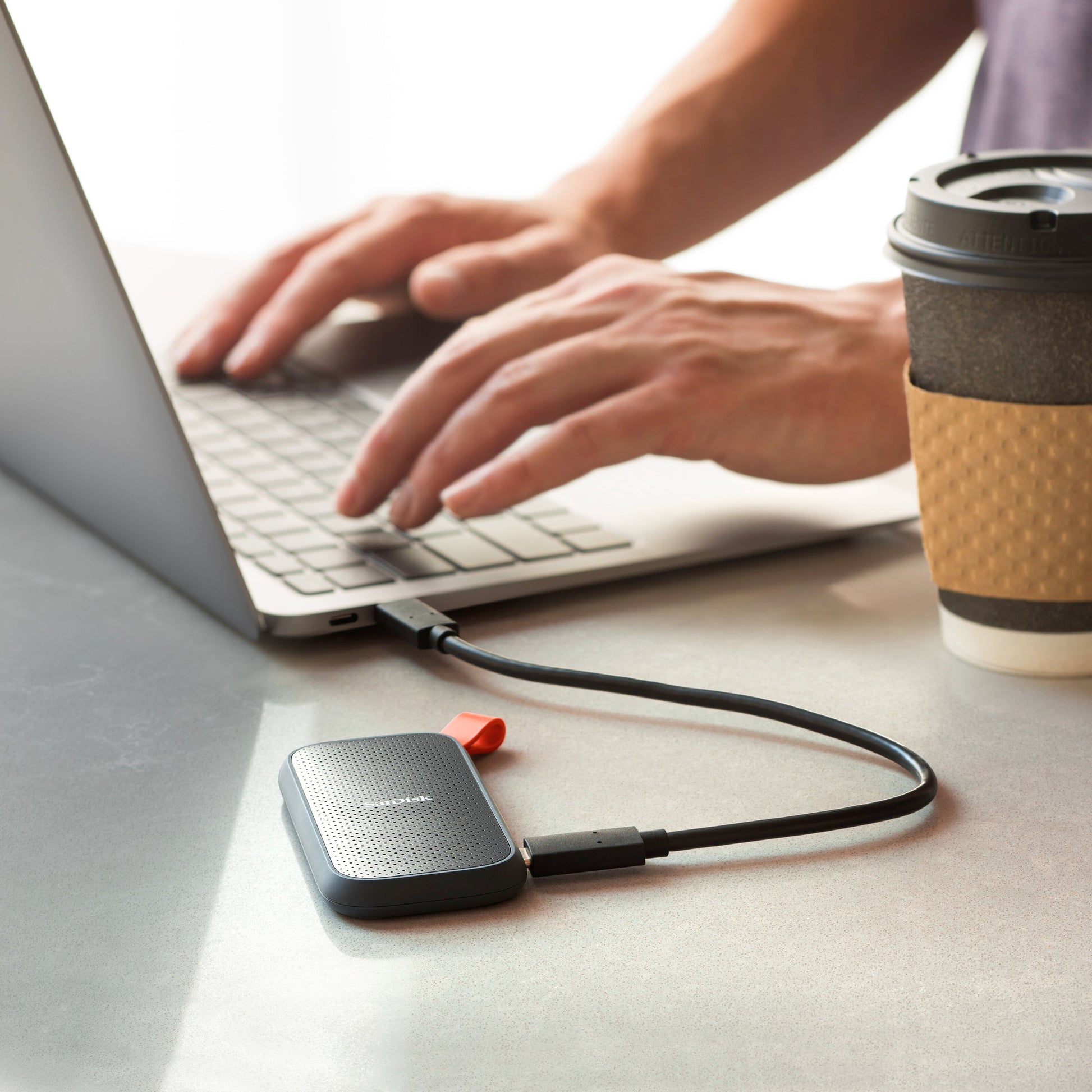 Sandisk bærbar SSD-harddisk 1TB tilsluttet via USB til laptop, synlig kaffekop på bordet.
