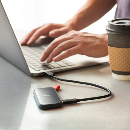 Sandisk bærbar SSD-harddisk 1TB tilsluttet via USB til laptop, synlig kaffekop på bordet.