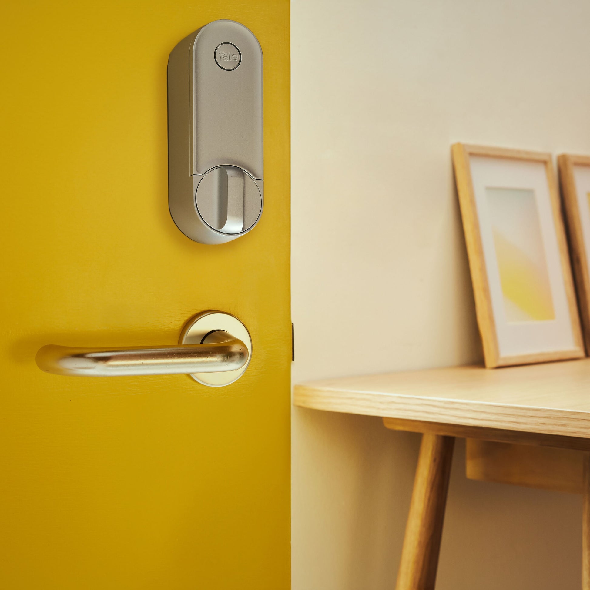 Yale Linus Smart Lock L2 Sølv monteret på gul dør, træbord med to indrammede billeder i baggrunden.