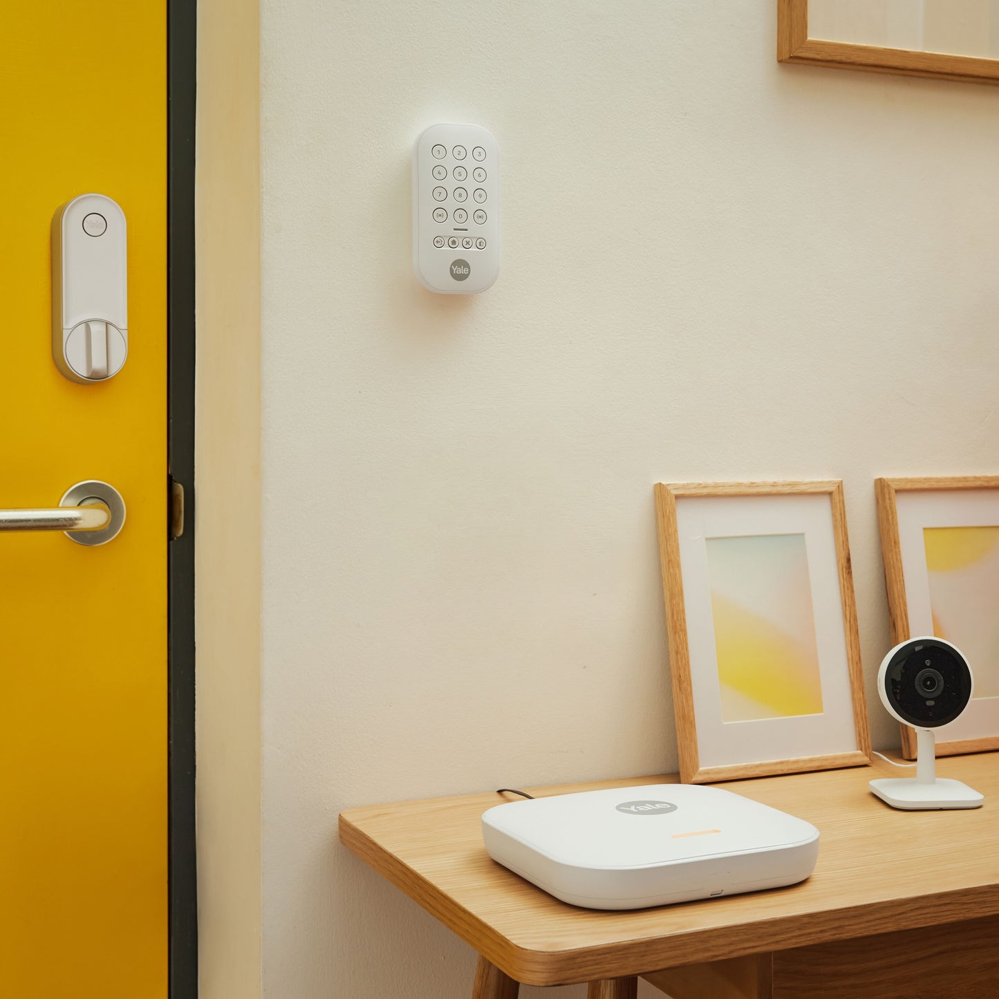 Yale Linus Smart Lock L2 sølv med tastatur på gul dør, synligt sikkerhedskamera og kontrolpanel på bord.