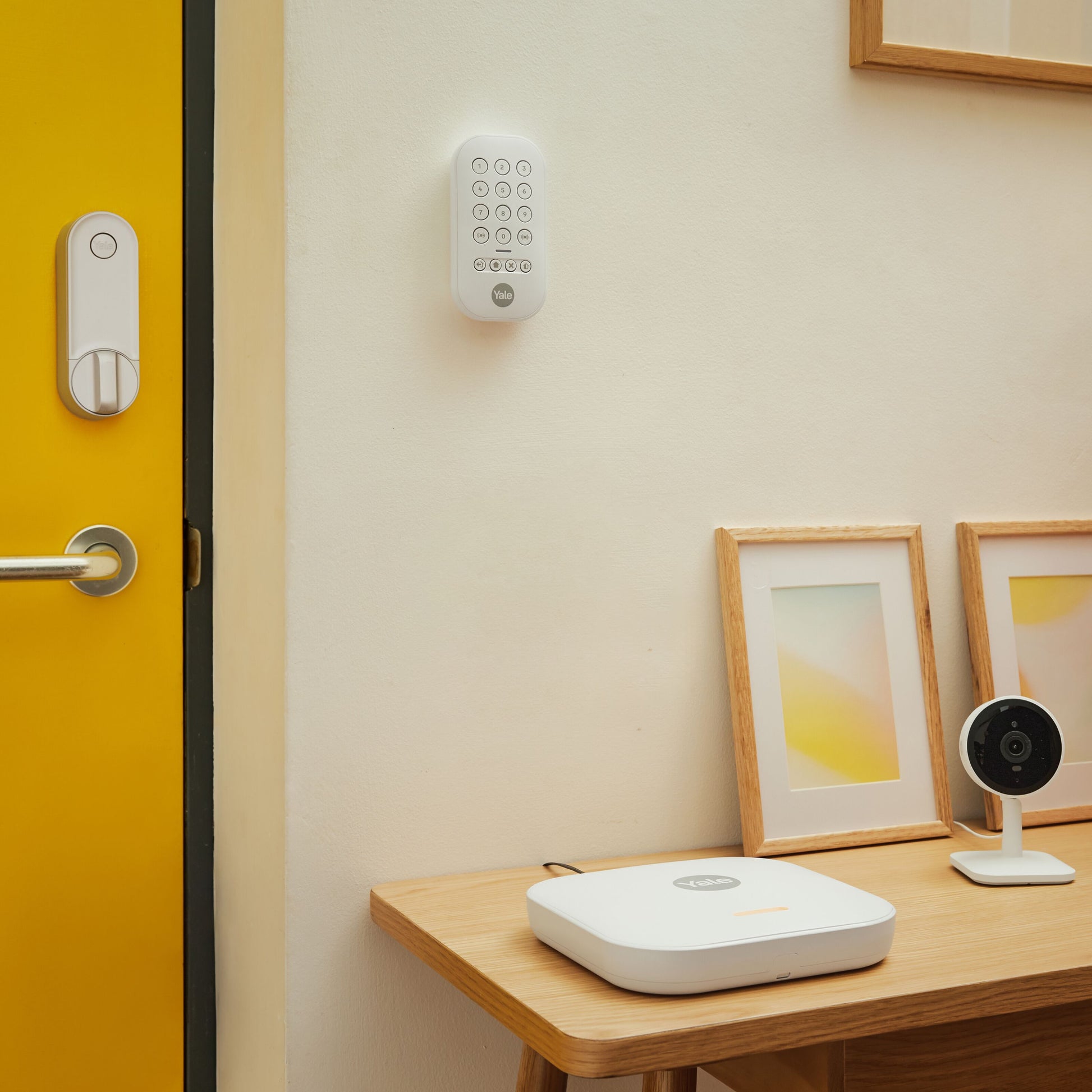 Yale Linus Smart Lock L2 sølv med tastatur på gul dør, synligt sikkerhedskamera og kontrolpanel på bord.