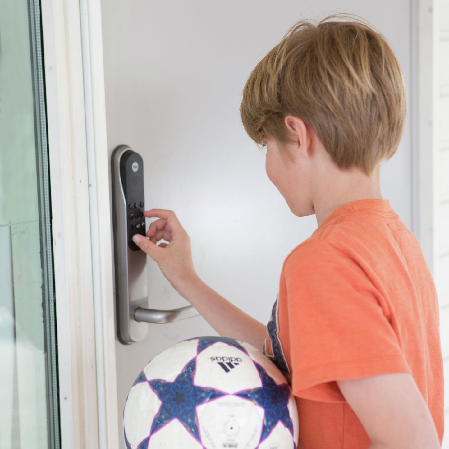 Yale Dørmand Klassisk Hjem elektronisk lås sølv, kodepanel, barn i orange trøje foran døren, fodbold i hånden.
