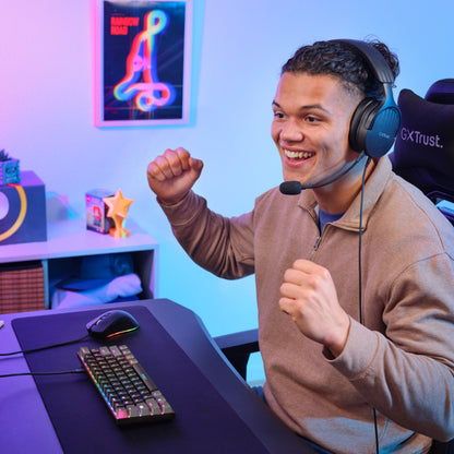 Person med headset ved skrivebord bruger Trust Acira GXT867 Tastatur 60 gaming chair og hylder i baggrunden.