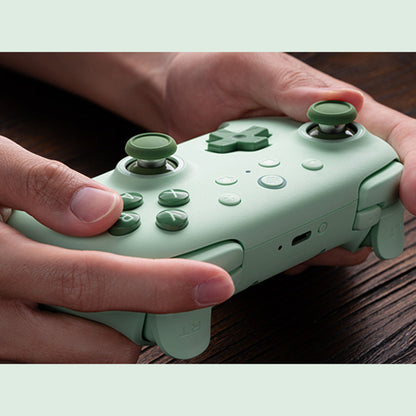 8Bitdo Ultimate 2C trådløs håndkontrol grøn Hall Effect holdes i begge hænder over mørkt træbord.