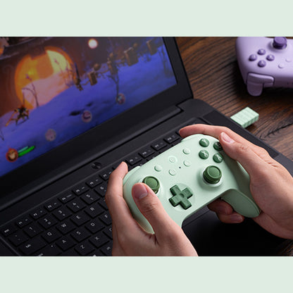 8Bitdo Ultimate 2C trådløs grøn håndkontrol med Hall Effect, lilla controller og USB-dongle på træbord ved laptop.