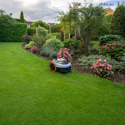Segway Navimow i105e robotplæneklipper 500 m² klipper græs ved lyserøde blomster omgivet af buske og træer.