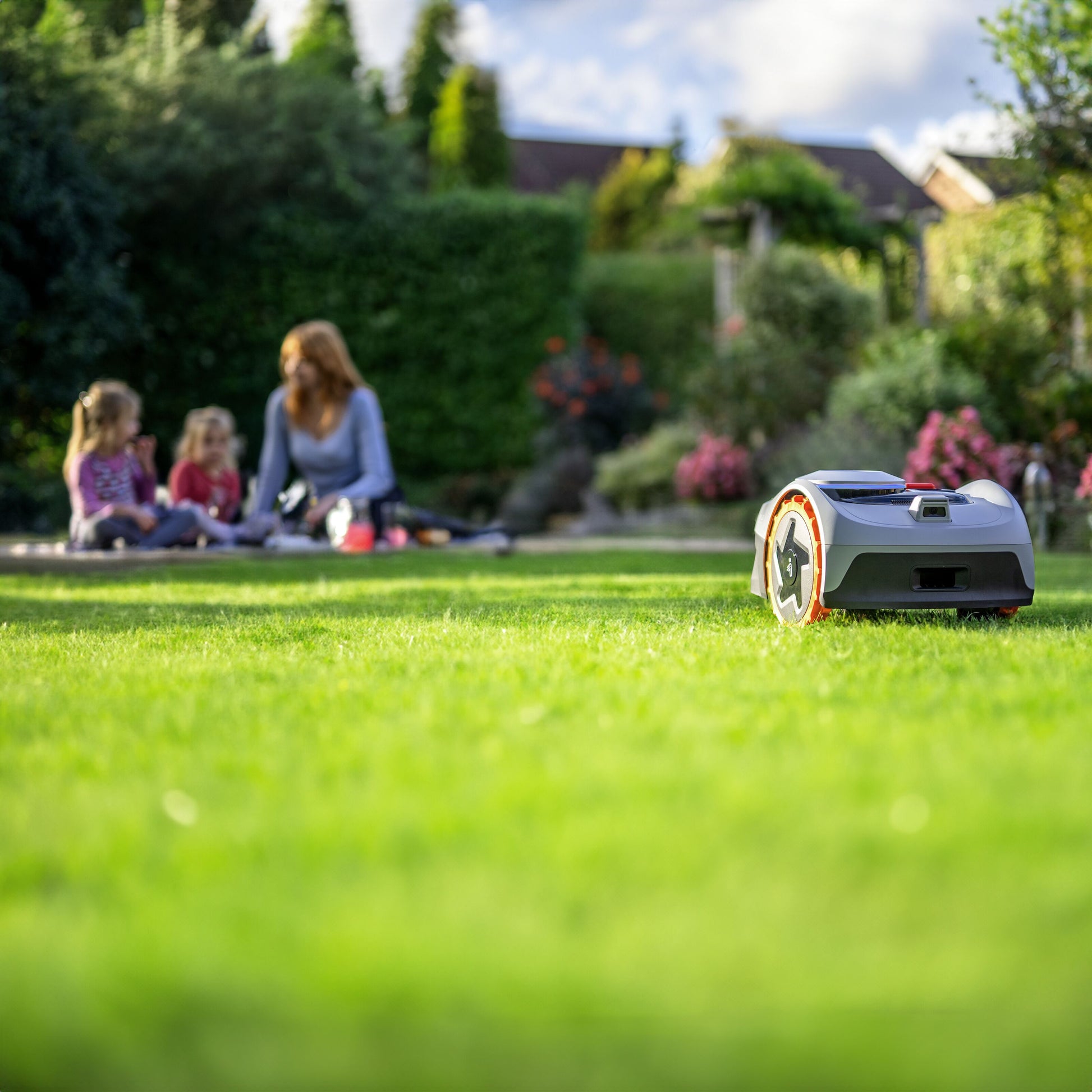 Segway Navimow i105e robotplæneklipper klipper græsset i forhaven, kvinde og to børn sidder på tæppe i baggrunden.