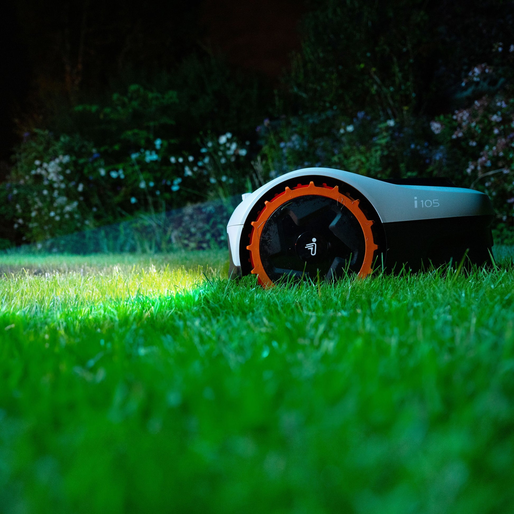 Segway Navimow i105e robotplæneklipper til 500 m² på græs omgivet af buske og hvide blomster ved skumring.