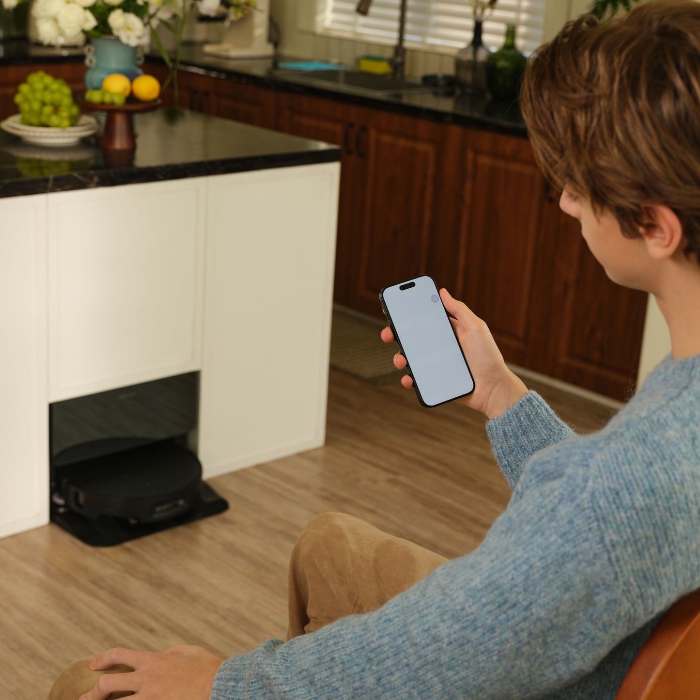 Roborock Saros 10 Sort robotstøvsuger under køkkenbord, person holder smartphone med blank skærm.