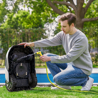 Dreame A1 Pro robotplæneklipper underside rengøres med gul haveslange udendørs, mand iført grå sweater og jeans.