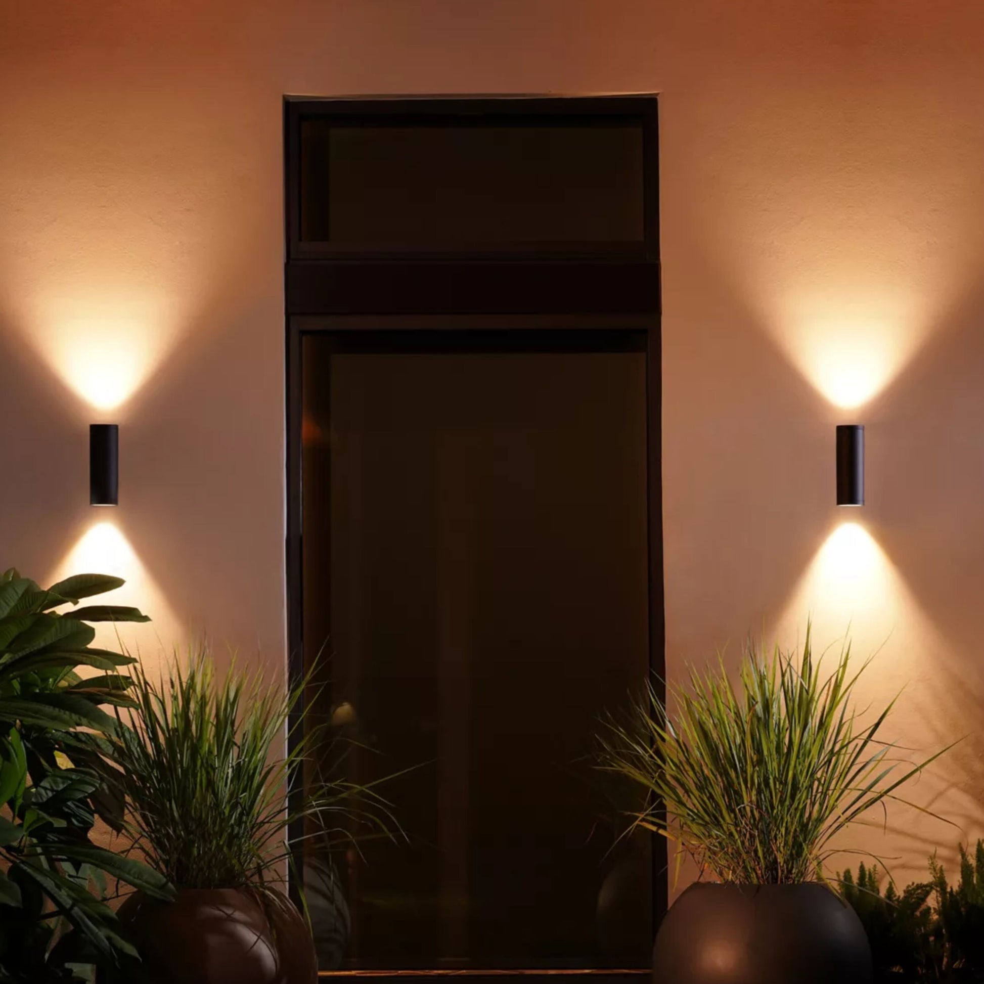 Philips Hue udendørs facadebelysning lyser op- og nedad ved indgang om natten, store krukker foran døren.