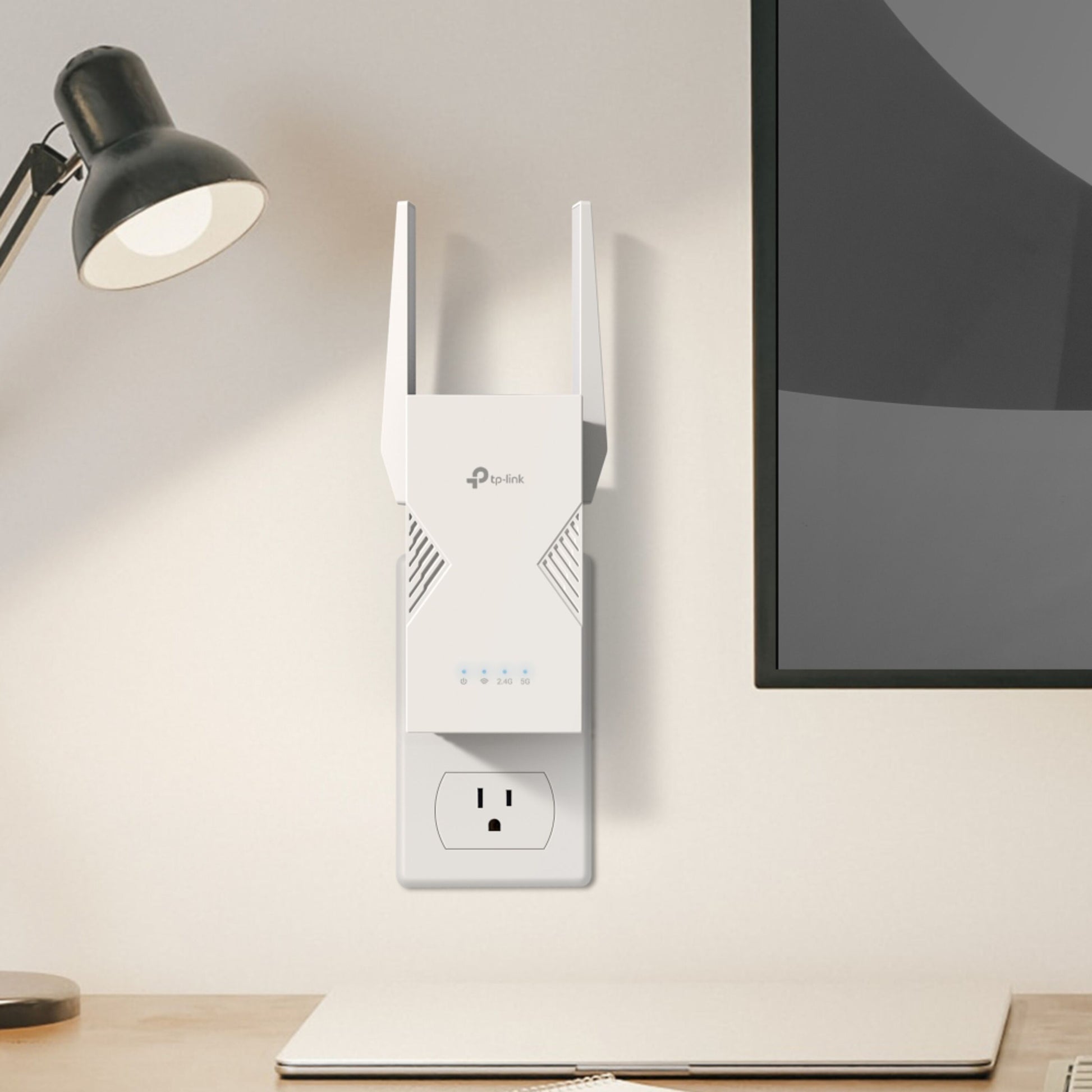 TP-Link RE235BE WiFi 7 BE3600-repeater i stikkontakt over skrivebord med lukket laptop, sort lampe og monitorskærm.