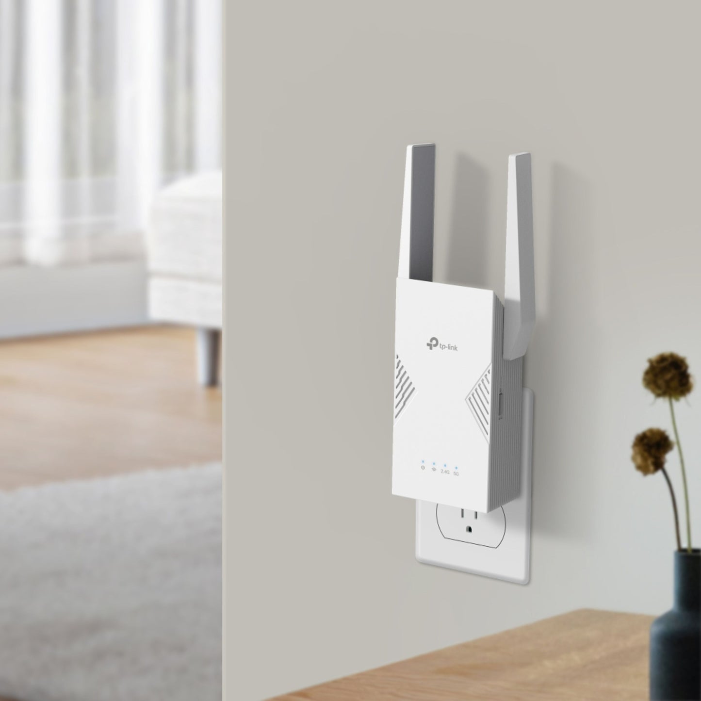 TP-Link RE235BE WiFi 7 BE3600 hvid dual-band repeater med to antenner i stikkontakt, bord med tørrede blomster ved siden af.