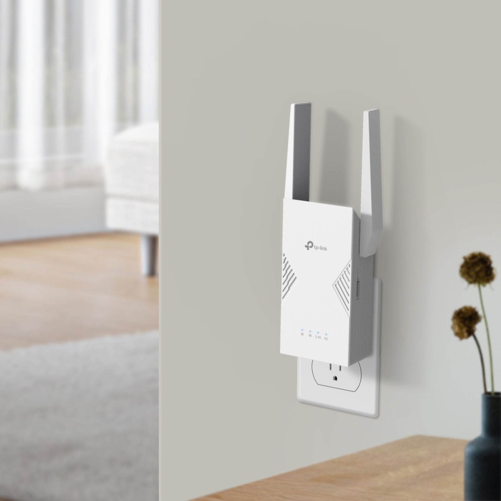 TP-Link RE235BE WiFi 7 BE3600 hvid dual-band repeater med to antenner i stikkontakt, bord med tørrede blomster ved siden af.