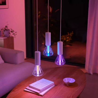 Philips Hue Lightguide Triangle E27 smart LED-pære 500 lm lyser i pink, lilla og blå i kolbeformet pendellampe over bord.