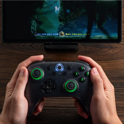 8Bitdo Ultimate 2C trådløs controller Transparent Sort, én i hånd foran skærm med spil, en anden på træoverflade.