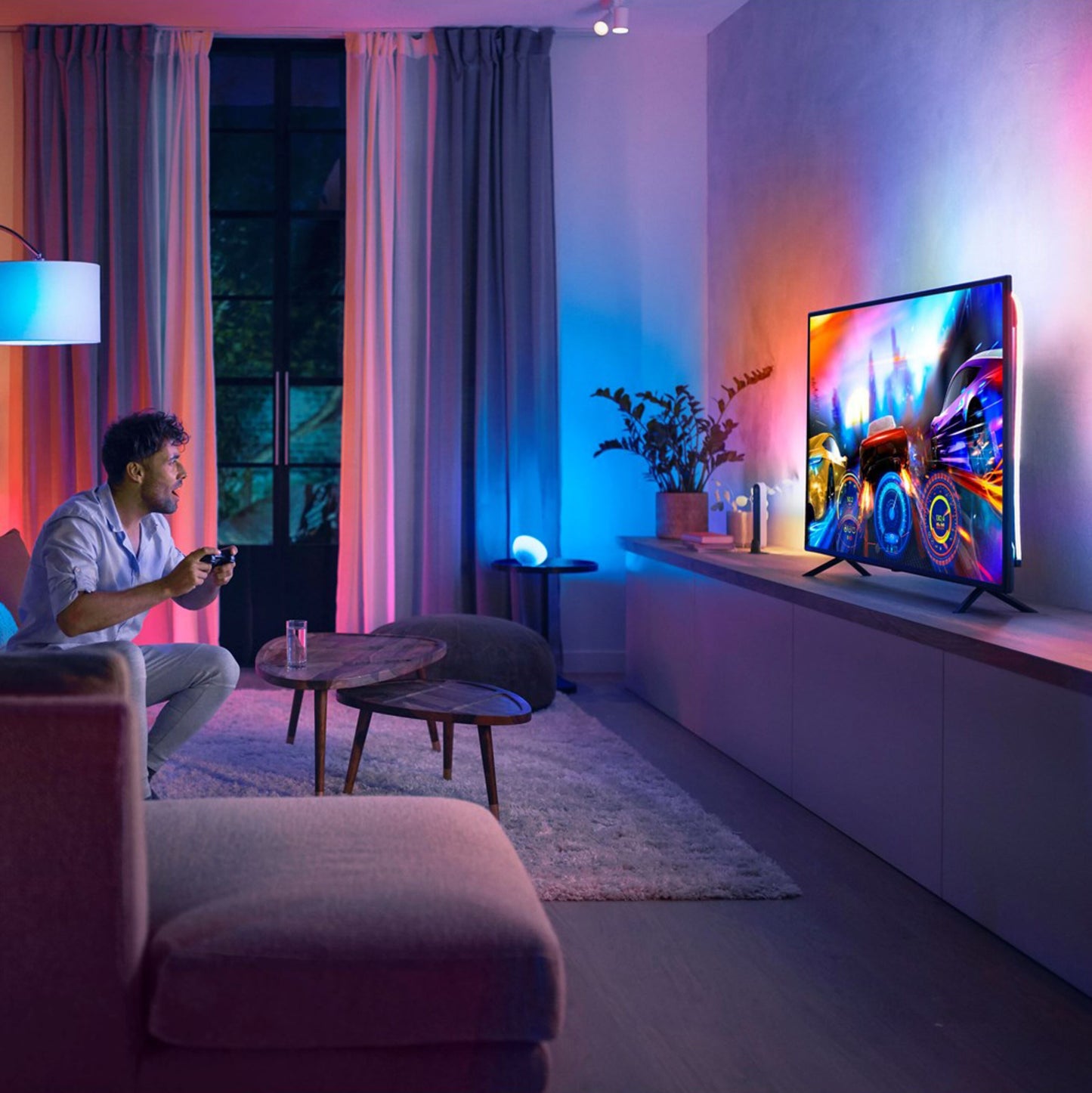 Philips Hue Play Gradient Lightstrip til TV 75", LED-lysliste monteret bag TV, sofabord og plante synlige.