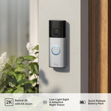 Ring Battery Video Doorbell Plus Retinal 2K i nikkel-sølv, blå lysende knap, monteret på væggen ved døren, ikon for 2K og nattesyn.