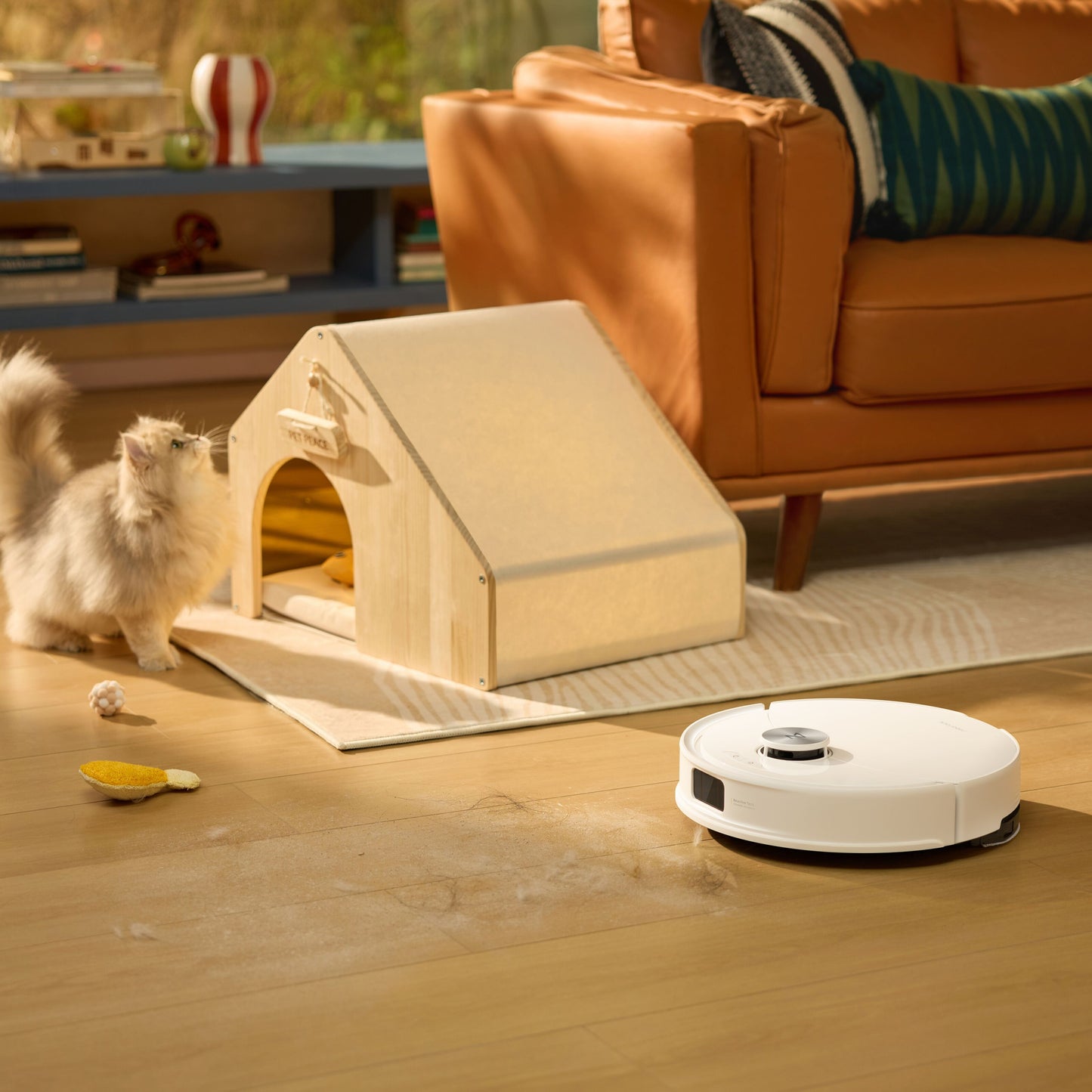 Roborock Q10 VF+ robotstøvsuger med ladestation hvid rengør trægulv nær kat og lille trædyrhus ved brun sofa.