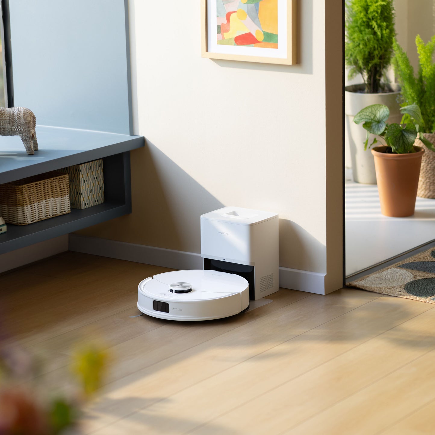 Roborock Q10 VF+ robotstøvsuger med ladestation på lyst trægulv, grønne planter og abstrakt vægkunst i baggrunden.