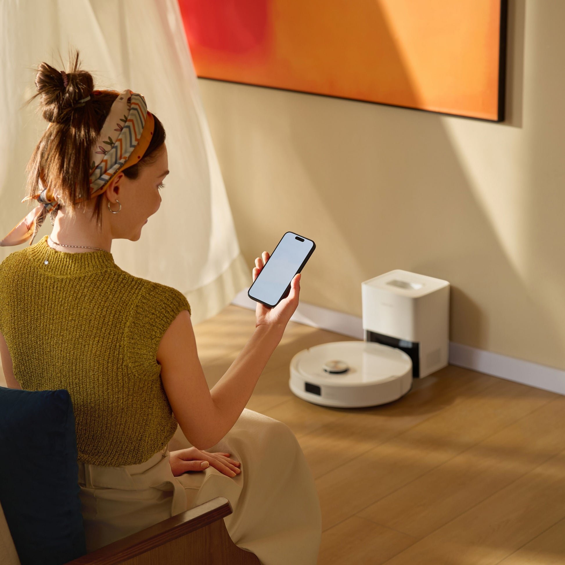 Roborock Q10 VF+ robotstøvsuger med ladestation hvid ved væg, kvinde i grønt ser på smartphone i forgrunden.