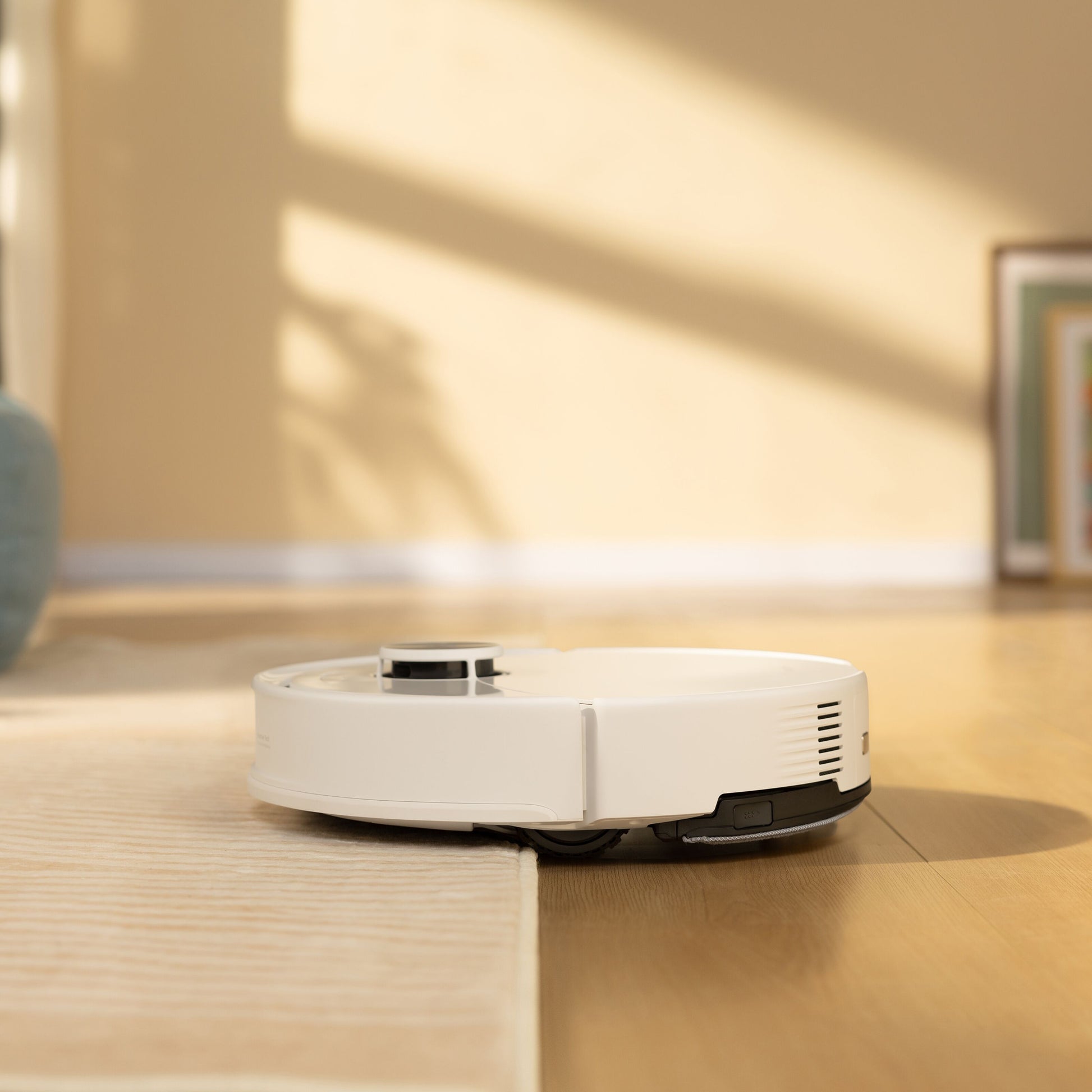 Roborock Q10 VF+ robotstøvsuger med ladestation på lyst trægulv i rum med beige vægge og indrammet billede.