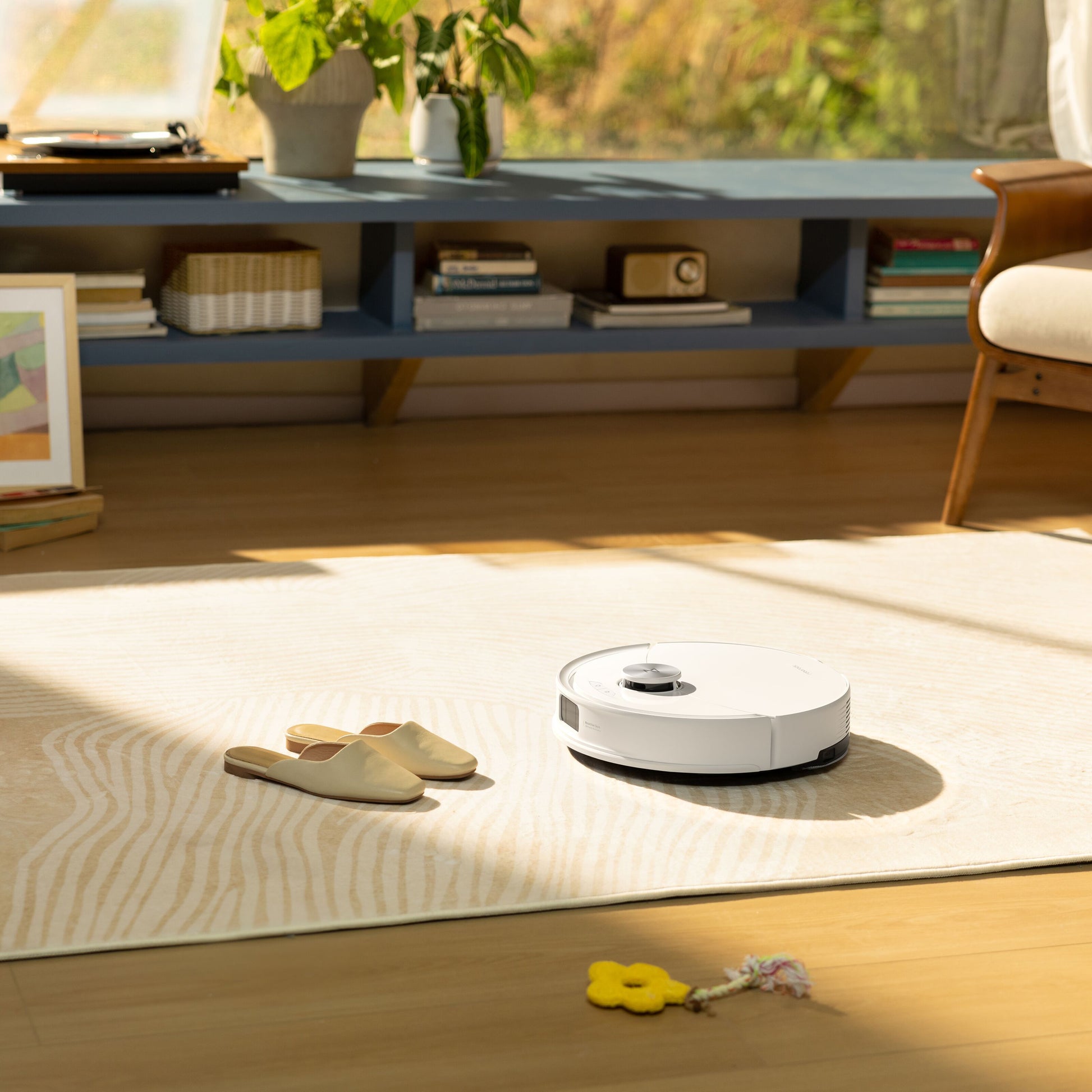 Roborock Q10 VF+ robotstøvsuger med ladestation på lyst tæppe, synlig beige hjemmesko og gult legetøj.