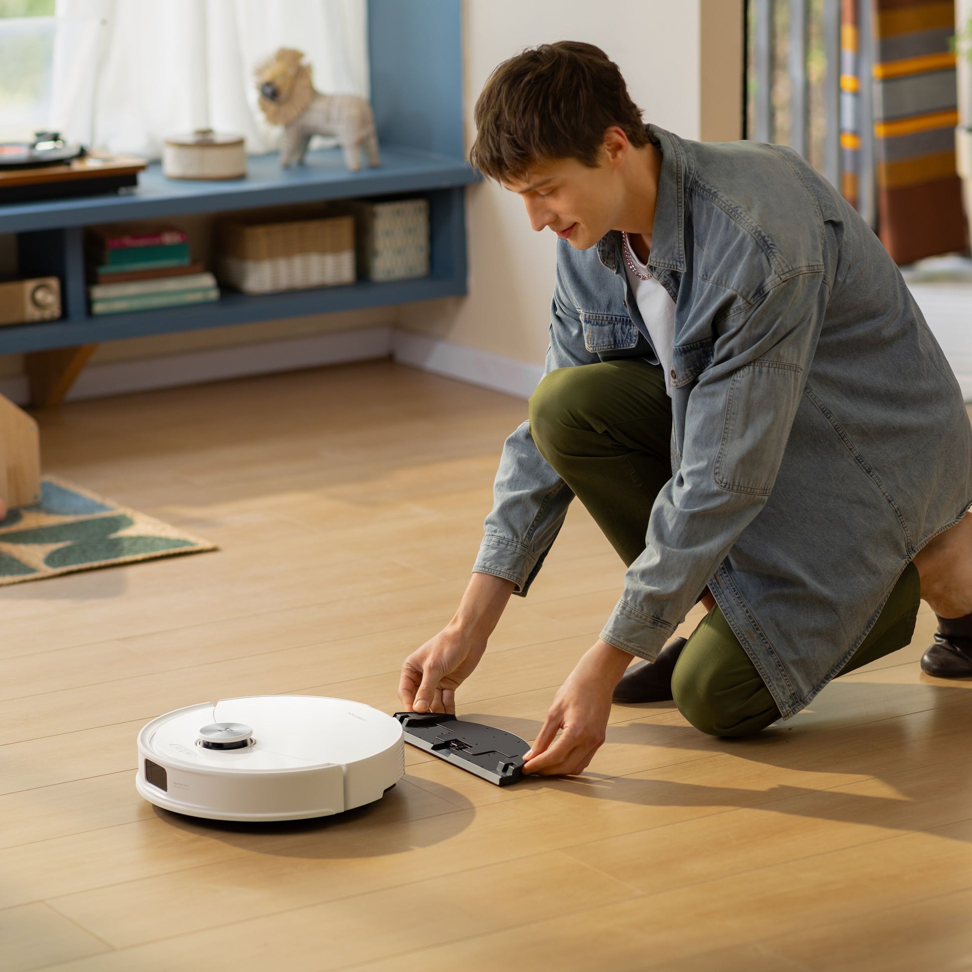 Roborock Q10 VF+ robotstøvsuger med ladestation hvid betjenes af person på trægulv i stue med reol, tæppe og vindue.