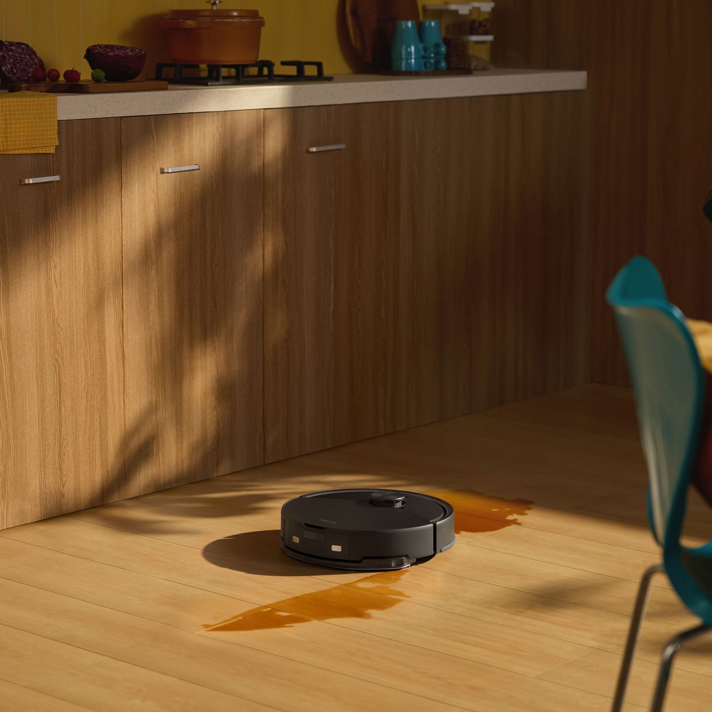 Roborock Q10 VF+ robotstøvsuger med ladestation fjerner orange spild på trægulv i køkken foran skabe og bordplade.