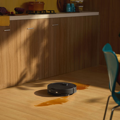 Roborock Q10 VF+ robotstøvsuger med ladestation fjerner orange spild på trægulv i køkken foran skabe og bordplade.