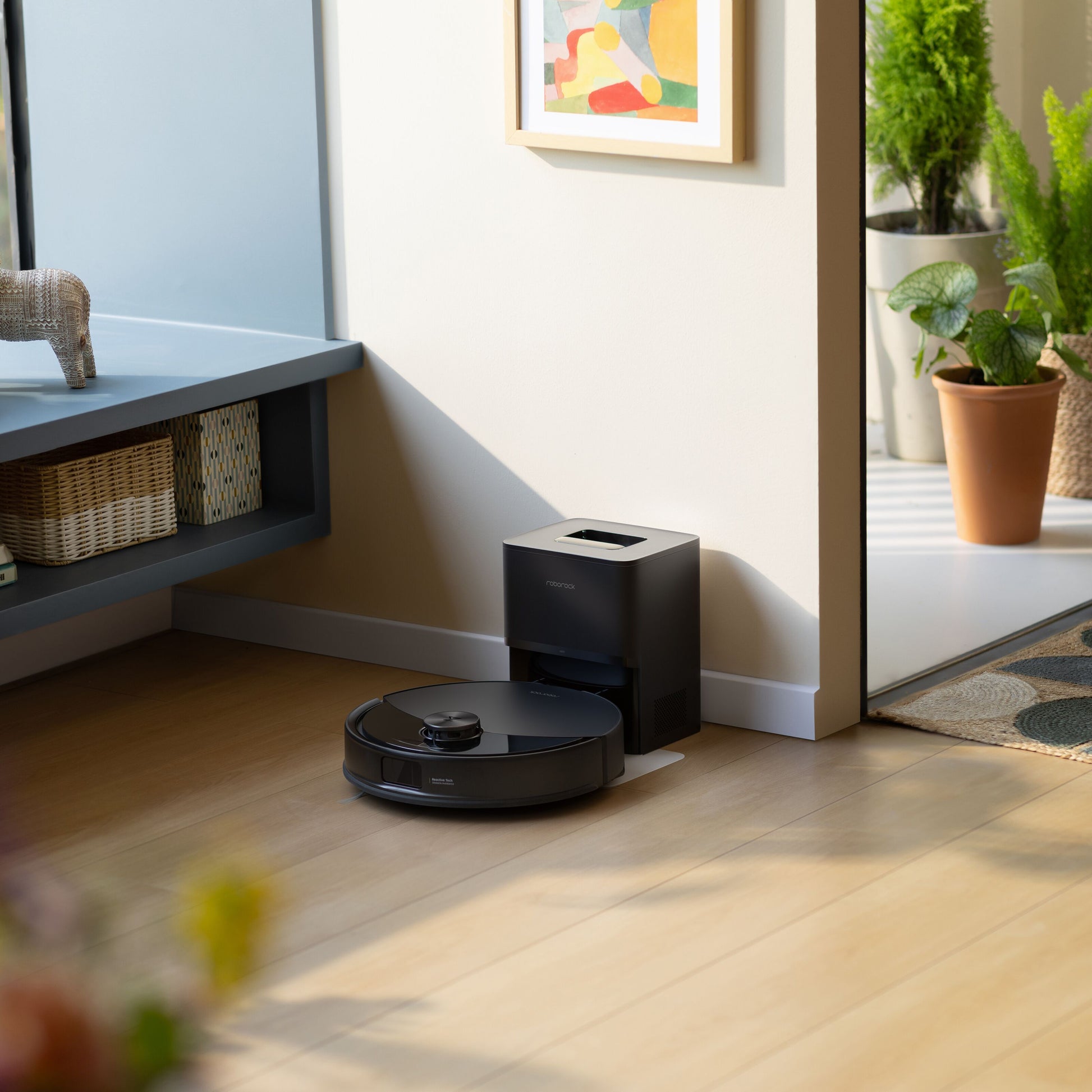Roborock Q10 VF+ robotstøvsuger sorterer ved ladestation på trægulv, grøn plante og abstrakt maleri synlige.