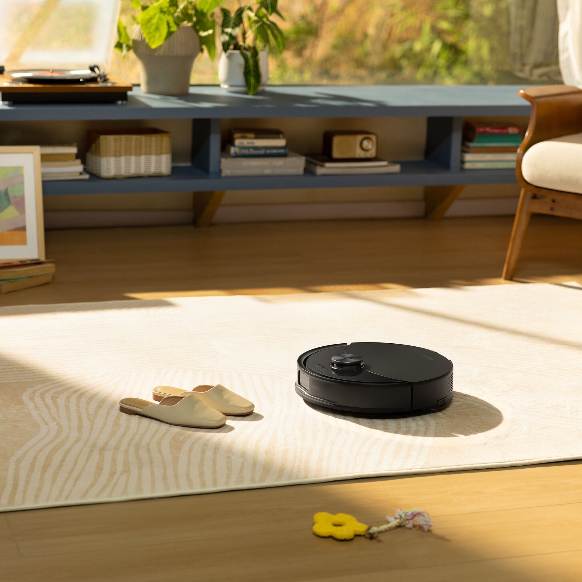 Roborock Q10 VF+ robotstøvsuger sort med ladestation kører på lyst mønstret tæppe ved hjemmesko og legetøj i stue.